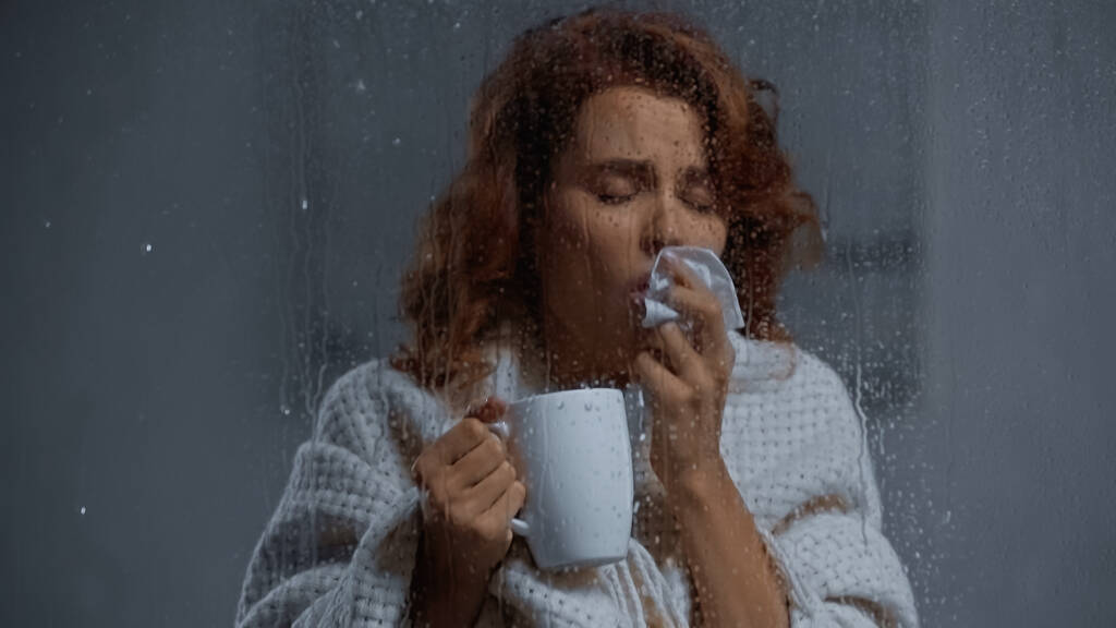 хвора жінка тримає чашку з гарячим напоєм і кашляє за віконним склом з краплями дощу
 - Фото, зображення