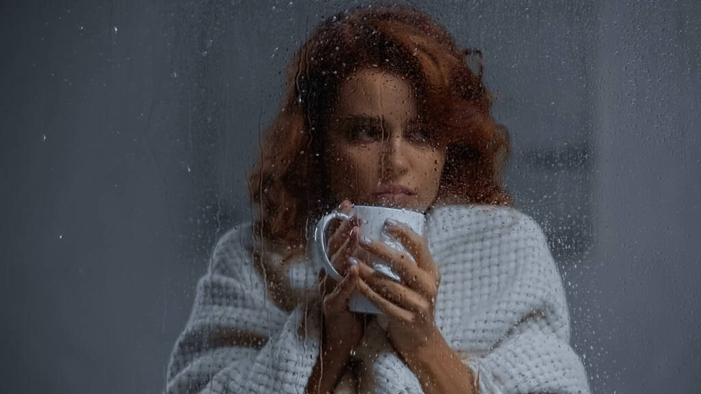 хвора жінка тримає чашку з гарячим напоєм за віконним склом з дощовими краплями
 - Фото, зображення