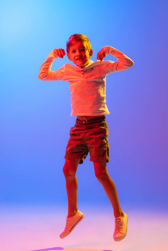 Springen. Studio shot van stijlvolle kleine jongen, kind poseren geïsoleerd op gradiënt blauw-paarse achtergrond in neon licht. Concept van emoties, gezichtsuitdrukking, mode, hobby 's, actieve levensstijl en reclame. - Foto, afbeelding