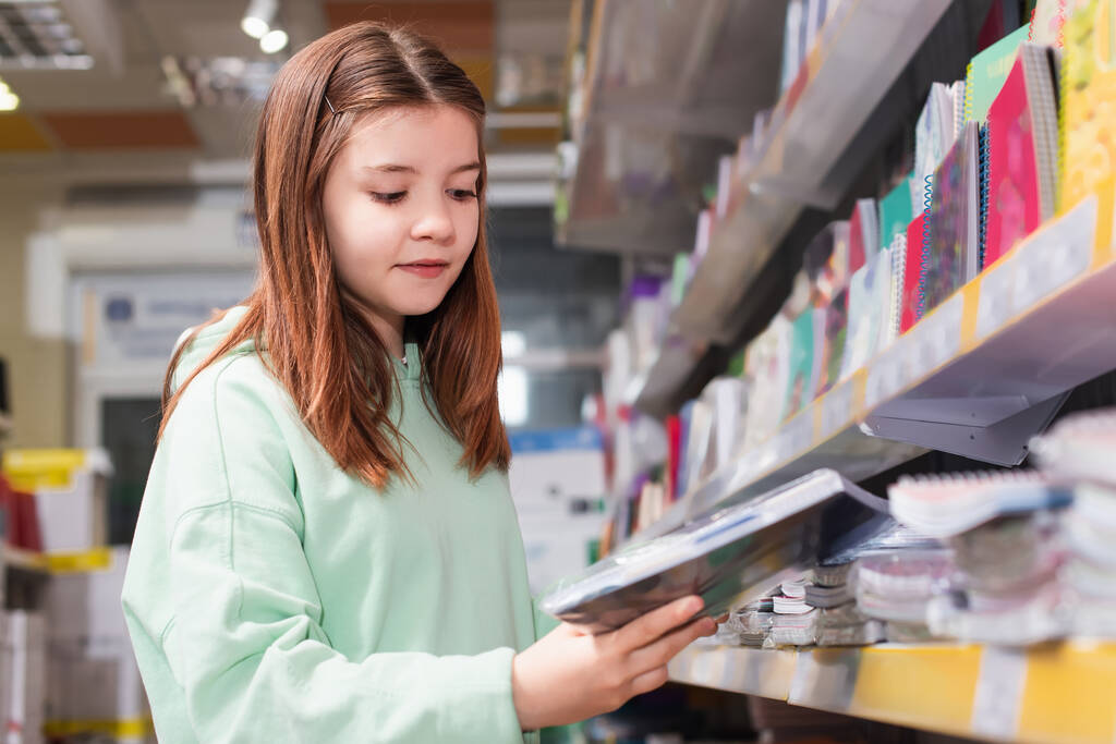 schoolgirl looking at new copybook near blurred rack in stationery shop - Zdjęcie, obraz