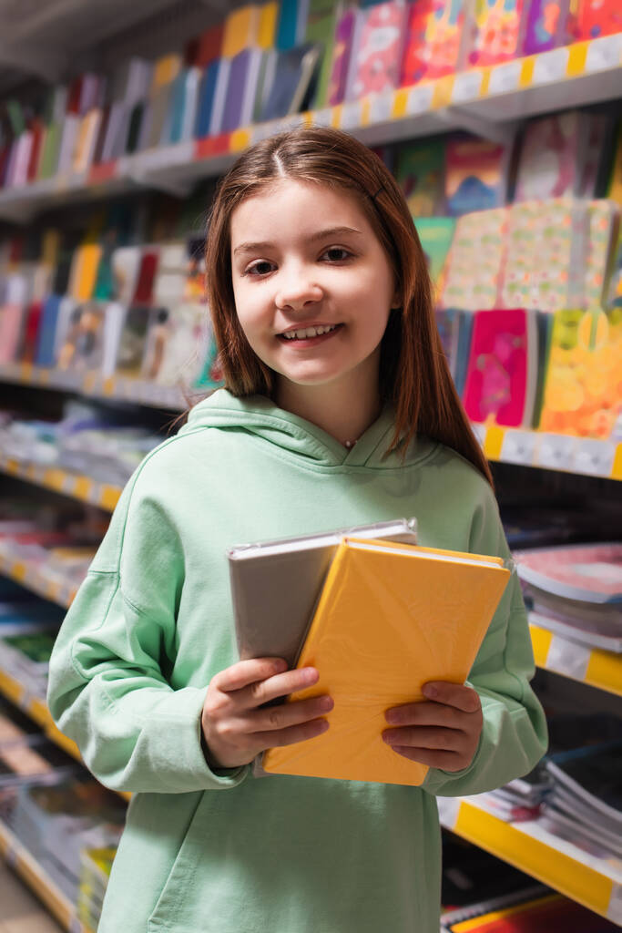 pleased girl with colorful notebooks smiling at camera in stationery store - Φωτογραφία, εικόνα