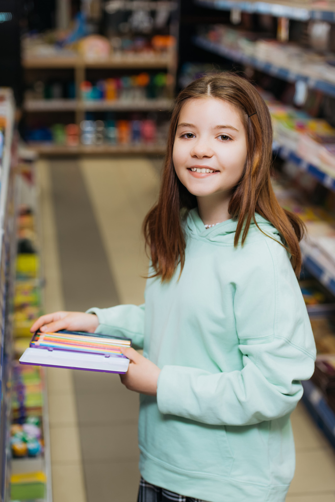 szczęśliwa dziewczyna z zestawem kolorowych ołówków patrząc na aparat fotograficzny w sklepie papierniczym - Zdjęcie, obraz