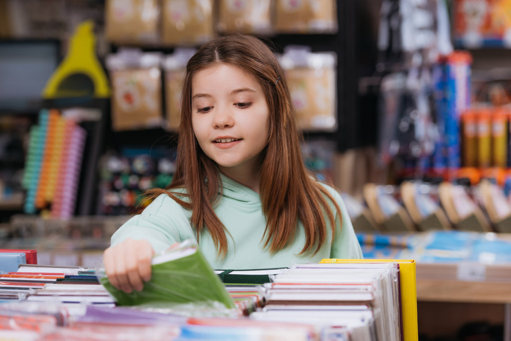 uśmiechnięta dziewczyna w pobliżu rozmazanych wielobarwnych książek do kopiowania w sklepie papierniczym - Zdjęcie, obraz