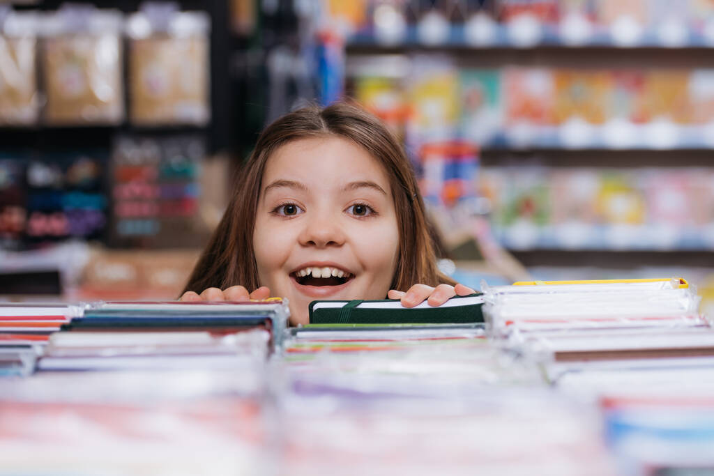 excited child looking at camera near blurred copybooks in stationery store - Photo, Image