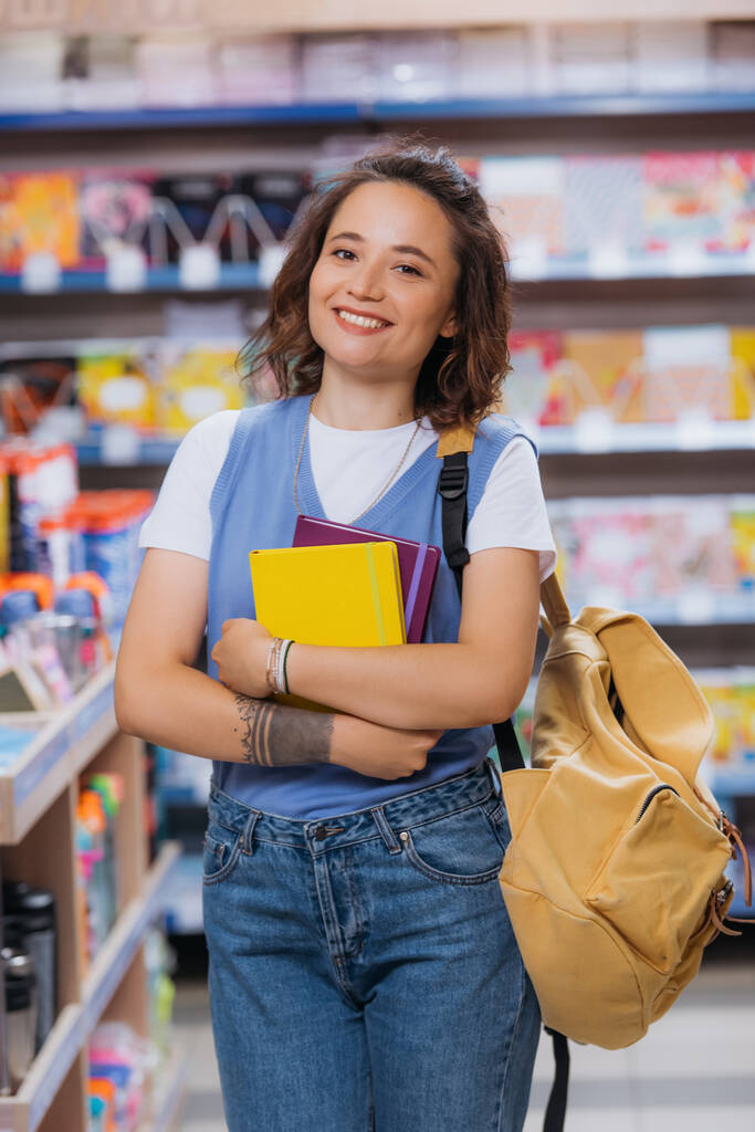 wytatuowana kobieta z nowymi notatnikami i plecakiem uśmiechnięta do kamery w sklepie papierniczym - Zdjęcie, obraz