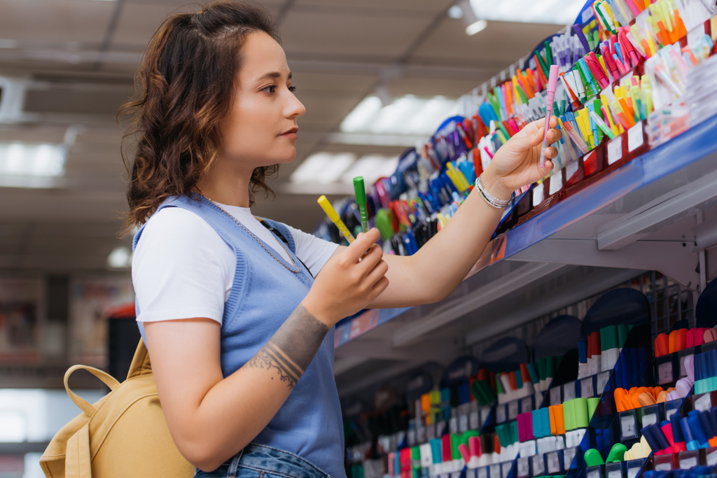 vista lateral da mulher tatuada segurando canetas multicoloridas perto do rack na loja - Foto, Imagem