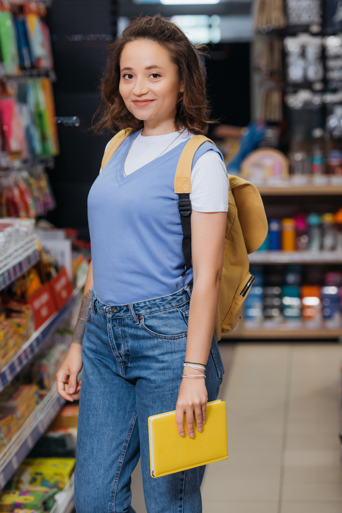 pleased woman with new copybook and backpack looking at camera in stationery shop - Fotoğraf, Görsel
