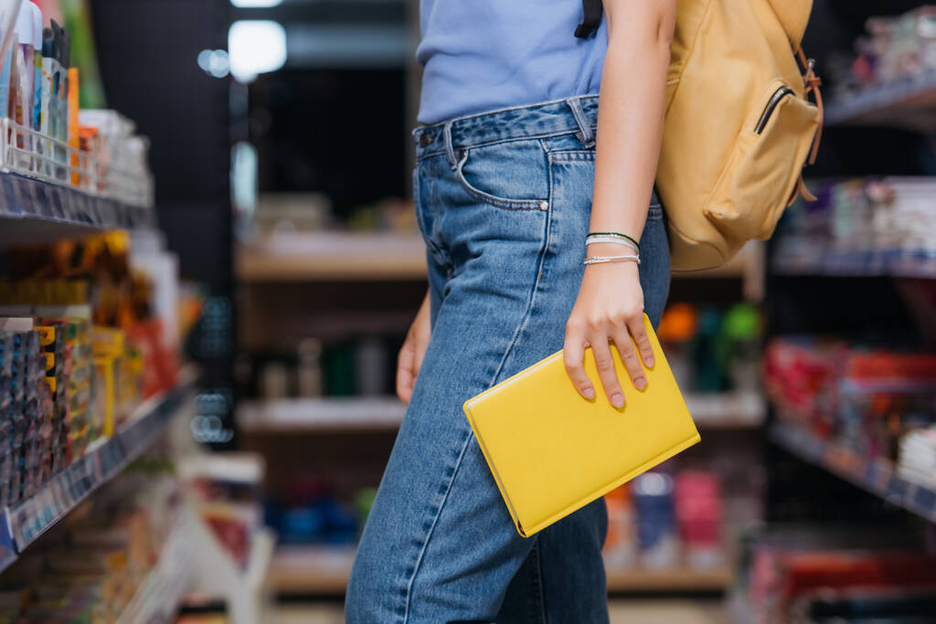 Ausgeschnittene Ansicht eines Studenten in Jeans und Perlenarmbändern, der ein neues Werbebuch im Laden hält - Foto, Bild