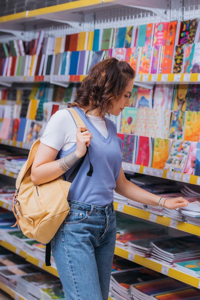 student met rugzak in de buurt rek met kleurrijke copybooks in briefpapier winkel - Foto, afbeelding