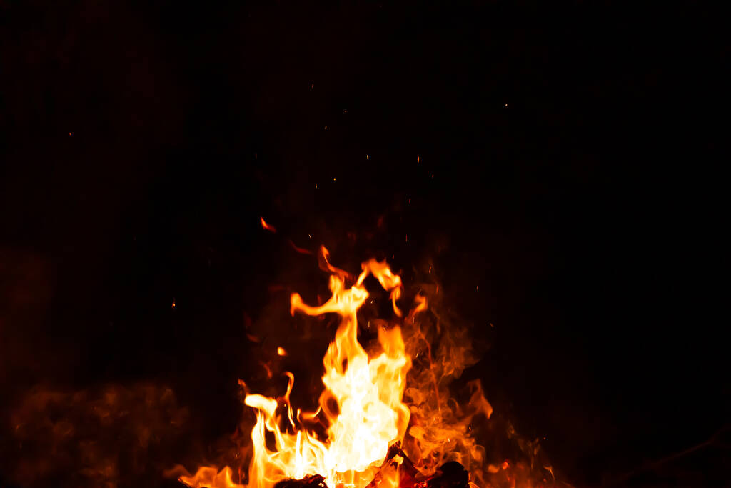 Горящие красные горячие искры поднимаются из большого огня ночью. Огонь искрится на заднем плане. Абстрактные темные блёстки огненных частиц. - Фото, изображение