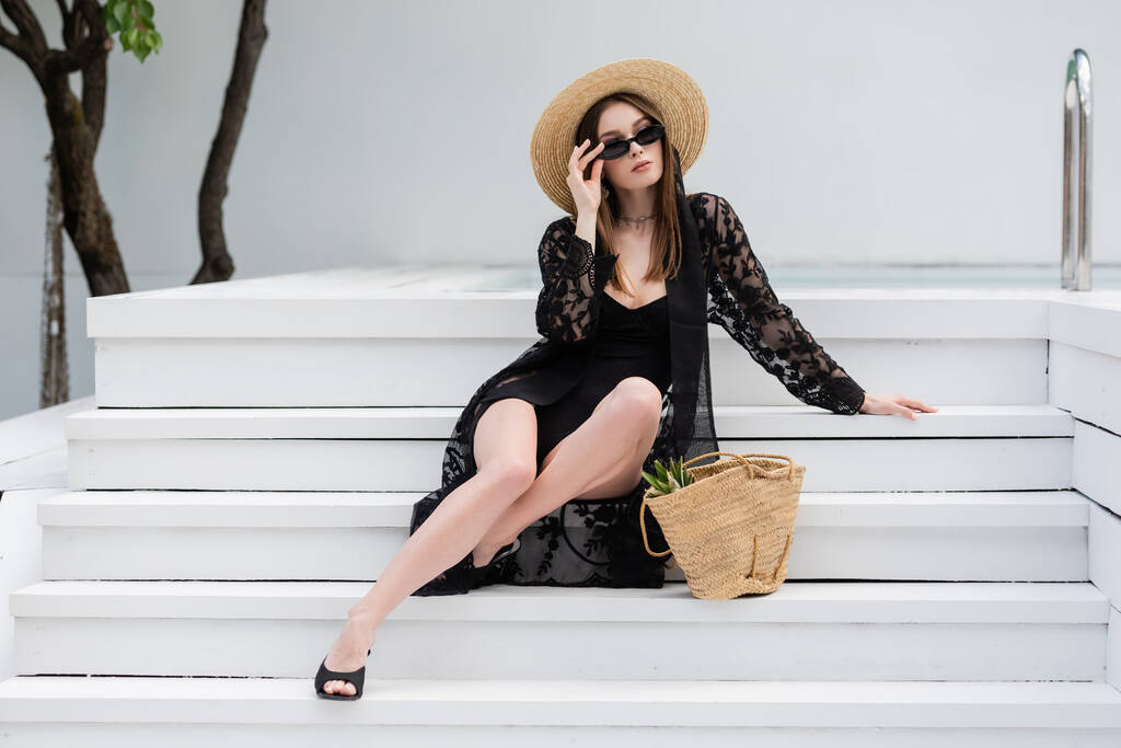 Fashionable woman in swimsuit and guipure robe holding sunglasses while sitting on stairs near pool  - Valokuva, kuva