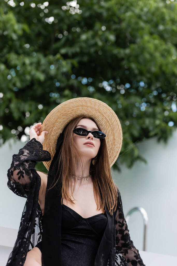 Fashionable woman in swimsuit and sunglasses holding straw hat at resort  - Photo, Image
