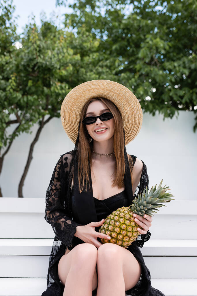 Cheerful woman in straw hat and swimsuit holding pineapple at resort  - Foto, imagen
