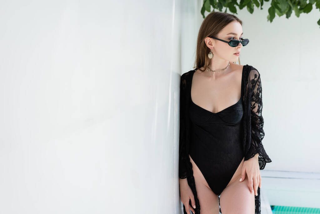 Stylish woman in sunglasses and swimsuit standing near wall at resort  - Fotó, kép