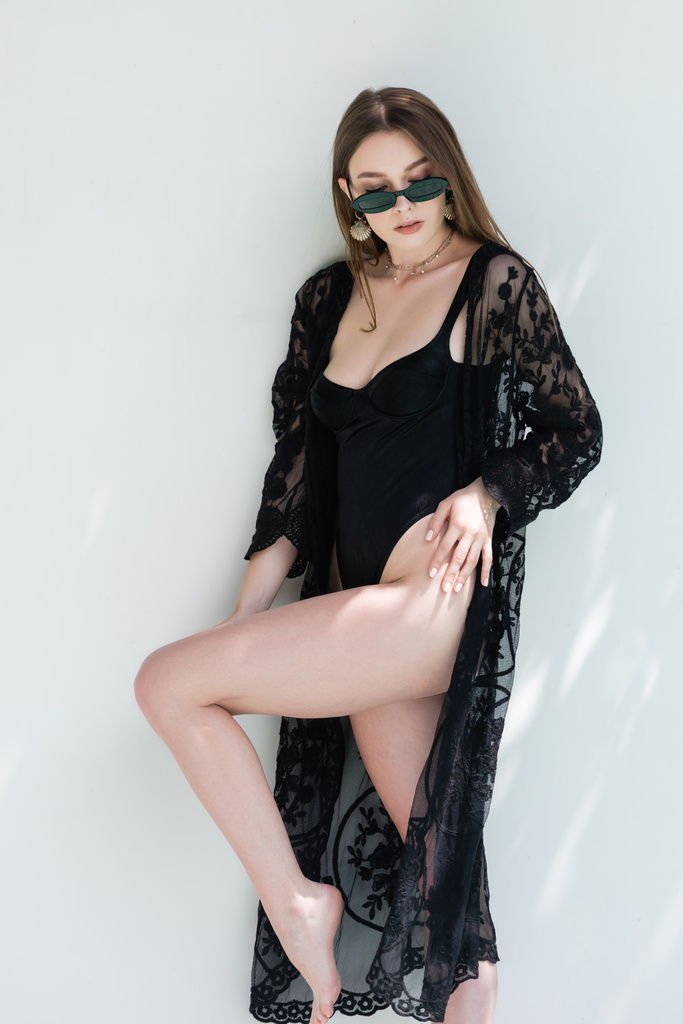 Stylish young woman in swimsuit and black guipure robe standing near wall  - Fotografie, Obrázek