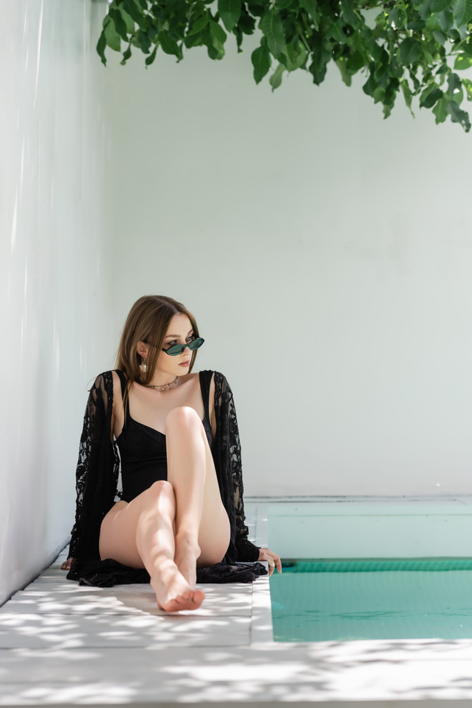 Modna kobieta w okularach przeciwsłonecznych i kostiumie kąpielowym z widokiem na basen w kurorcie  - Zdjęcie, obraz