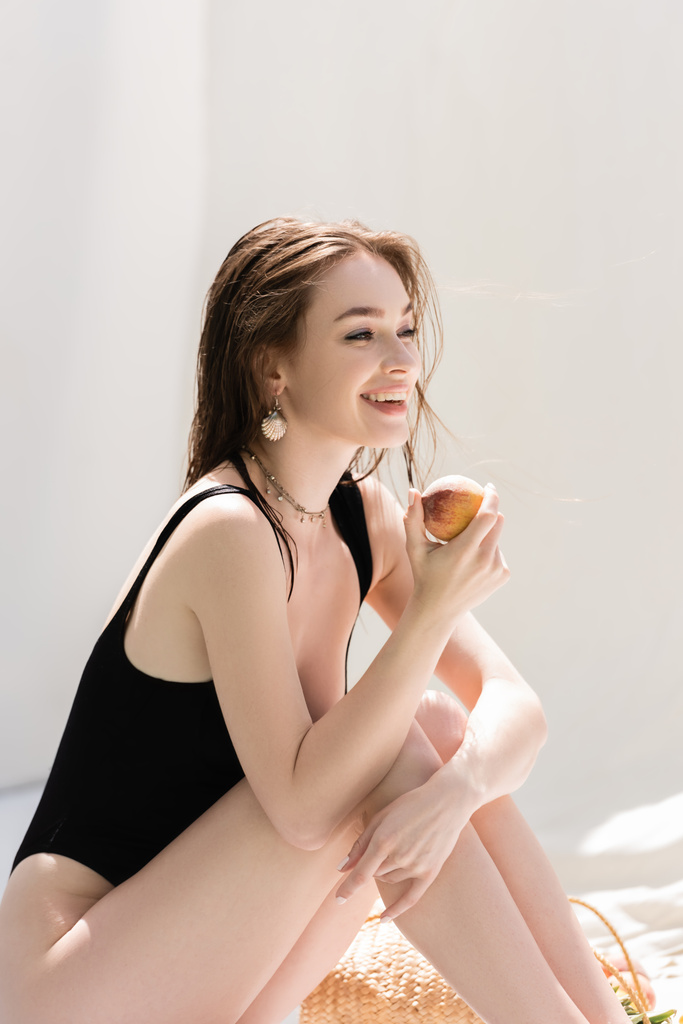Positive young woman in swimsuit holding peach near handbag at resort  - Φωτογραφία, εικόνα