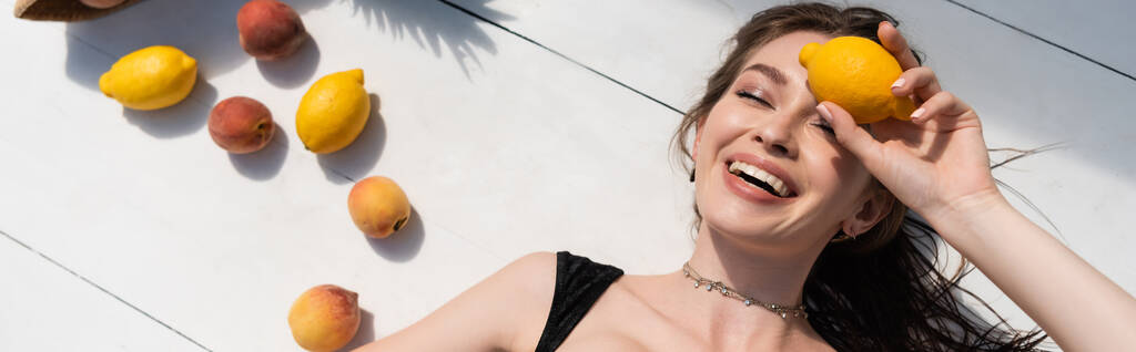 vista superior da jovem mulher feliz em maiô preto deitado perto de frutas frescas na superfície branca, banner - Foto, Imagem