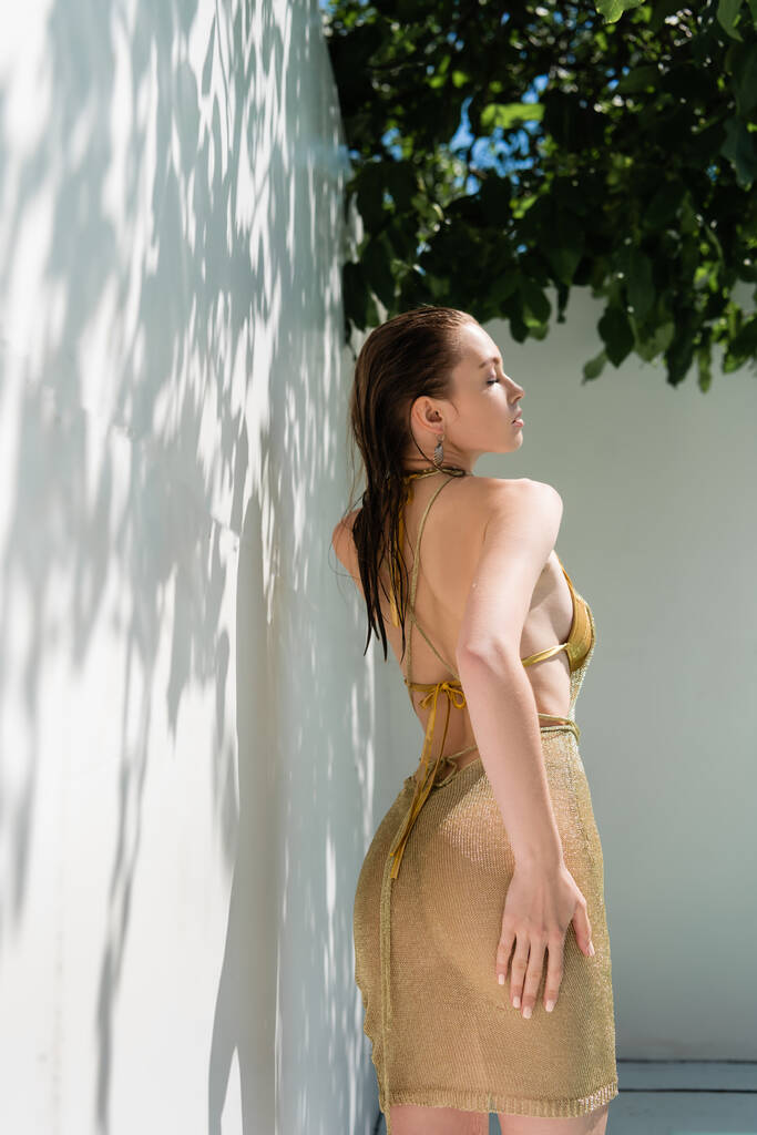 спокуслива жінка в золотій літній сукні, що стоїть біля стіни з тінями від зеленого листя
 - Фото, зображення
