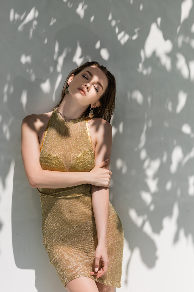 hezká mladá žena se zavřenýma očima pózující v letních šatech v blízkosti bílé zdi se stíny z listí  - Fotografie, Obrázek