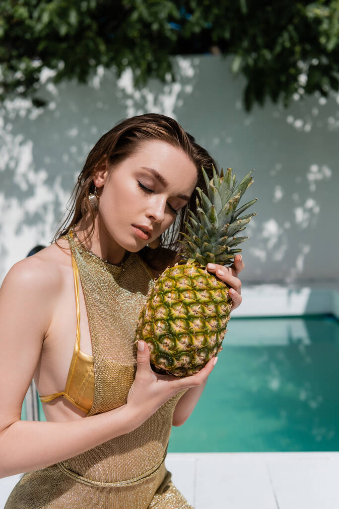 jovem mulher com olhos fechados em vestido de verão sentado com abacaxi na beira da piscina - Foto, Imagem