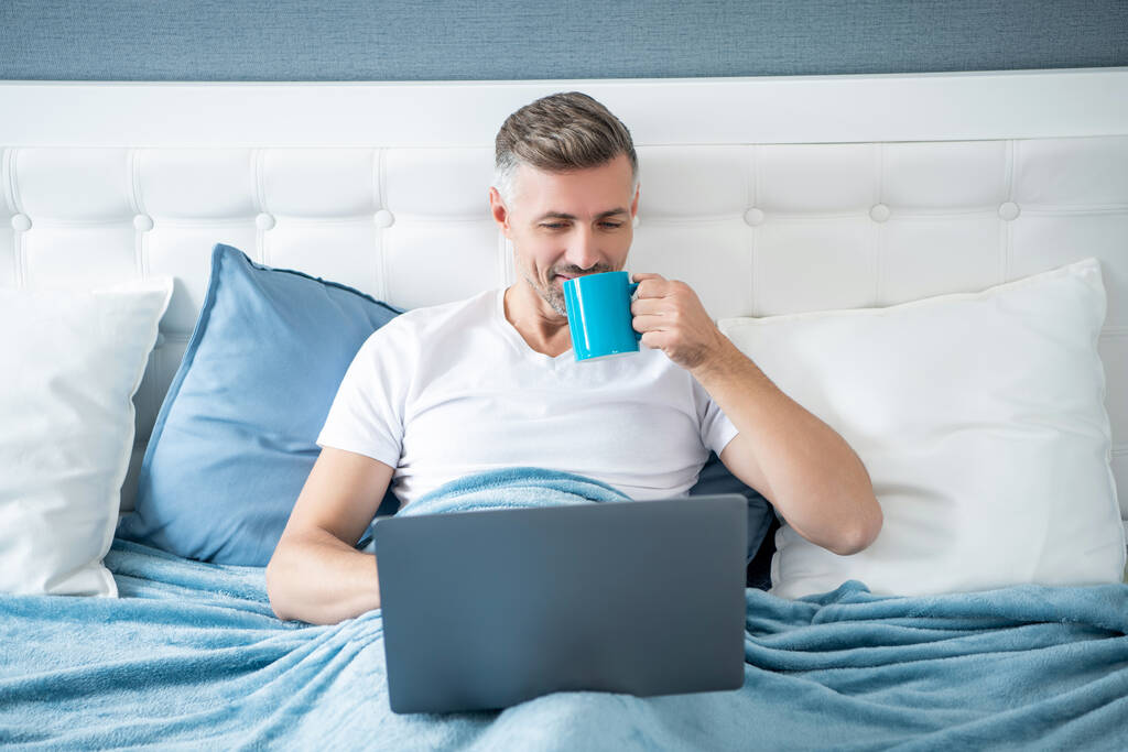 allegro uomo maturo che lavora sul computer portatile a letto con il caffè. - Foto, immagini