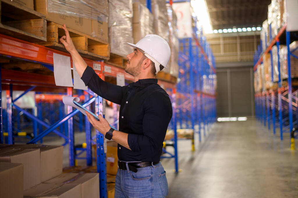 Young worker wearing helmet checking inventory and counting product on shelf in modern warehouse. - Foto, Imagen