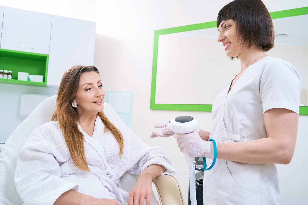 Usmívající se kosmetička informující pacientku o zákroku fotorejuvenace kůže - Fotografie, Obrázek
