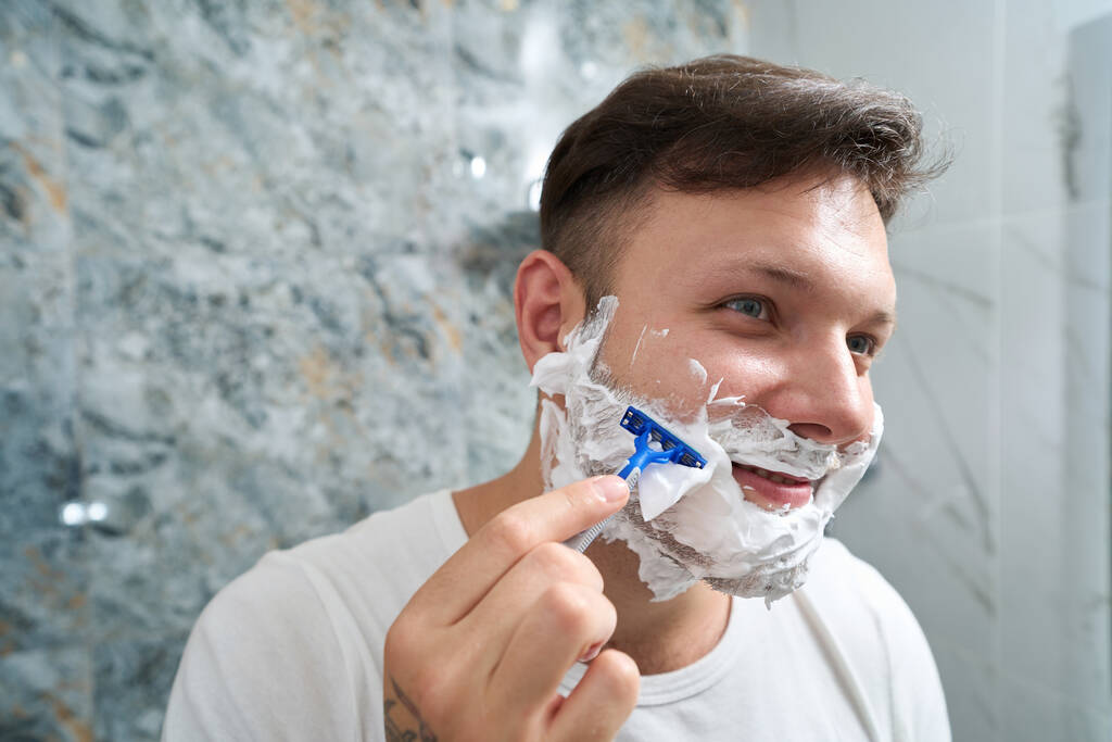Young man shaves in the bathroom, he is in a good mood, smile on face, shaving foam on cheeks - Photo, Image