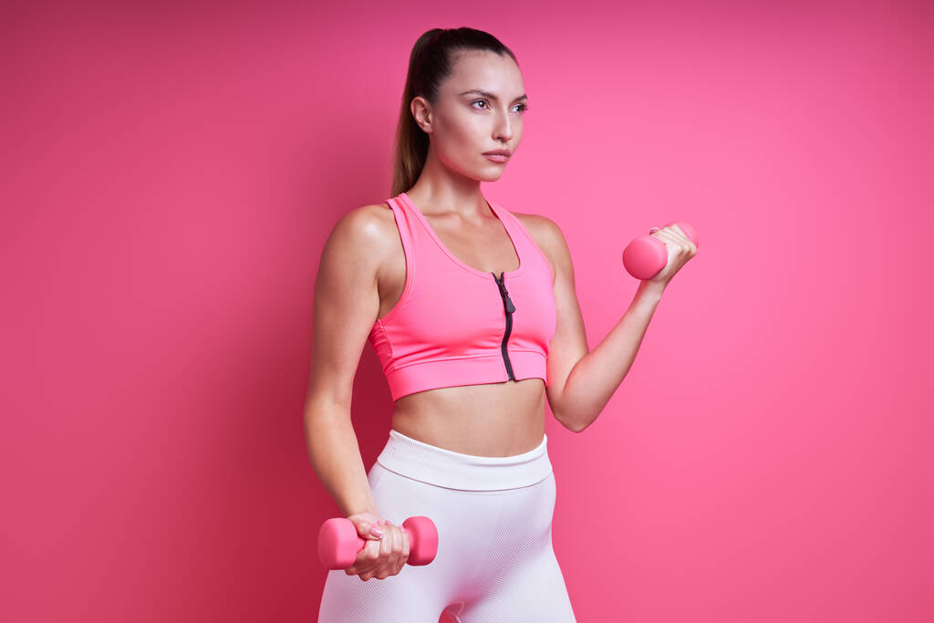 Confiant jeune femme en vêtements de sport exercice avec haltères sur fond rose - Photo, image