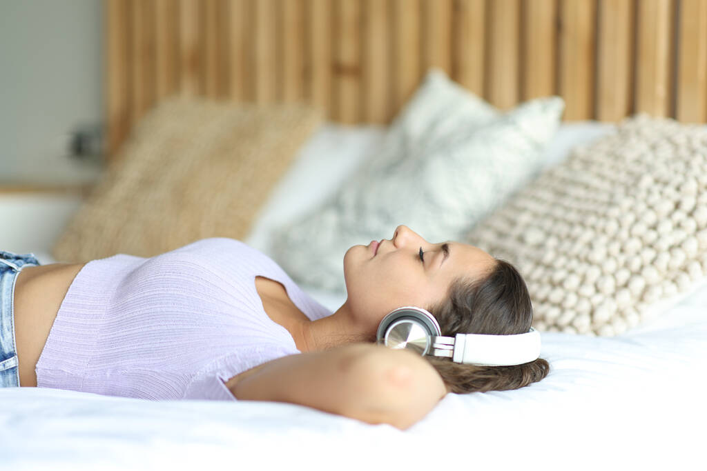 Портрет жінки в бездротових навушниках, що розслабляють слухати музику на ліжку в кімнаті
 - Фото, зображення