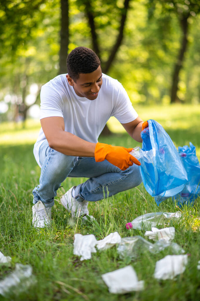 Social position. Smiling dark-skinned man in gloves crouching putting plastic in bag picking up trash in forest - Photo, Image