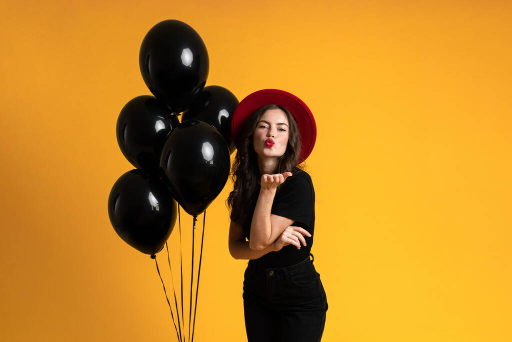 White woman blowing air kiss while posing with black balloons isolated over yellow background - 写真・画像
