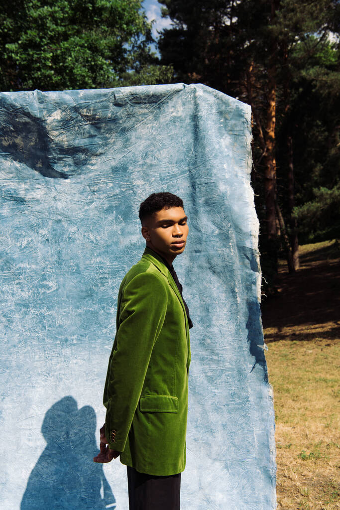 african american man in green blazer posing with closed eyes and hands behind back near blue drape - Foto, Imagen