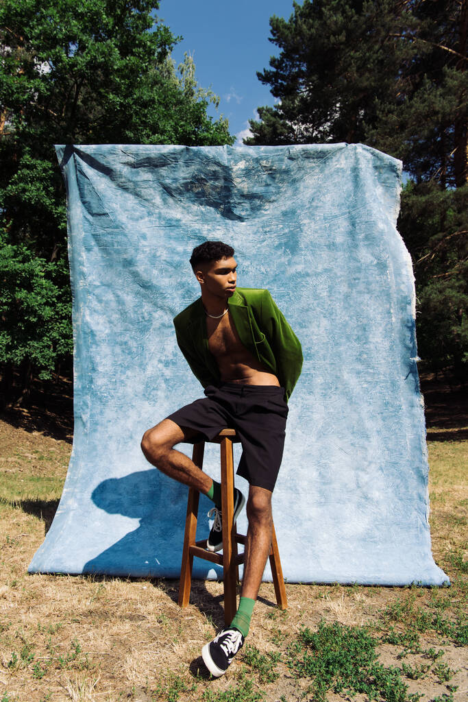 african american man in green blazer and back shorts posing on stool near blue drape in forest - Foto, Imagem