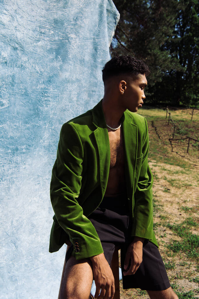 Yeşil kadife ceketli ve siyah şortlu genç Afro-Amerikan adam mavi kumaşın yanında ormanda oturuyor. - Fotoğraf, Görsel