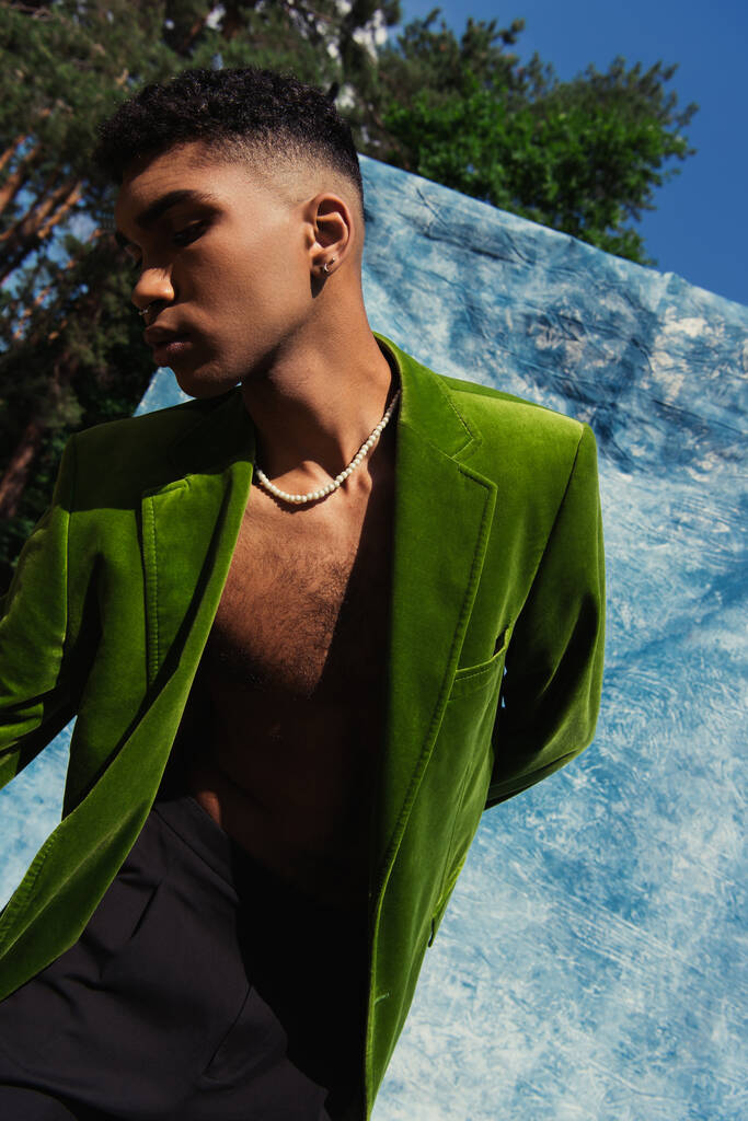modny Afroamerykanin człowiek pozowanie w zielony marynarka w pobliżu niebieski tkanina w parku - Zdjęcie, obraz