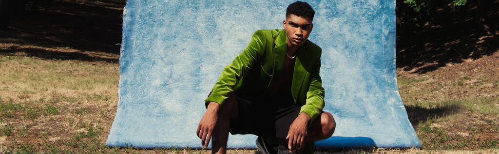 young african american man in green velvet blazer posing near blue textured cloth outdoors, banner - 写真・画像