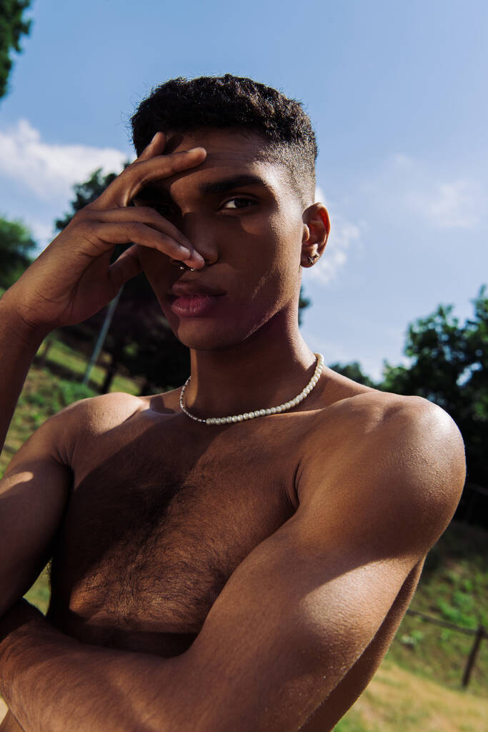 young shirtless african american man obscuring face with hand while looking at camera outdoors - Foto, Imagem