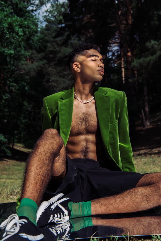 jonge en stijlvolle Afro-Amerikaanse man in groene blazer en zwarte shorts zittend op spiegel oppervlak in het bos - Foto, afbeelding