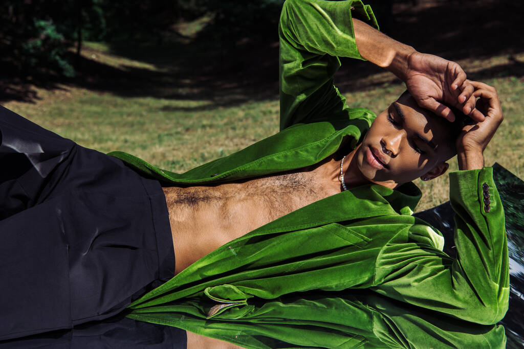 nuori afrikkalainen amerikkalainen mies trendikäs vihreä bleiseri makaa kädet lähellä päätä heijastava pinta puistossa - Valokuva, kuva
