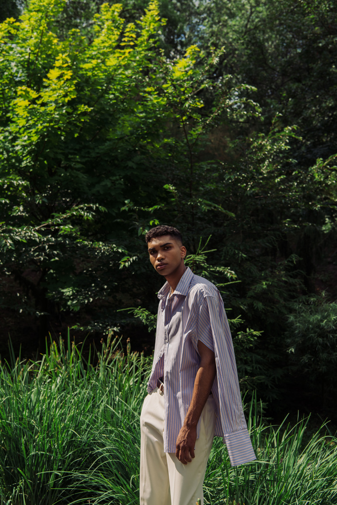 fiatal afro-amerikai férfi kék és csíkos ing nézi kamera a nyári parkban - Fotó, kép