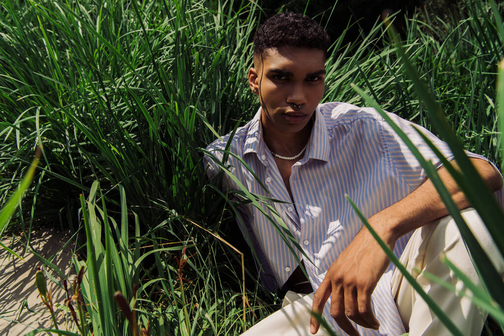 hoge hoek uitzicht van jonge Afrikaan amerikaanse man in trendy shirt zitten op gras en kijken naar camera - Foto, afbeelding