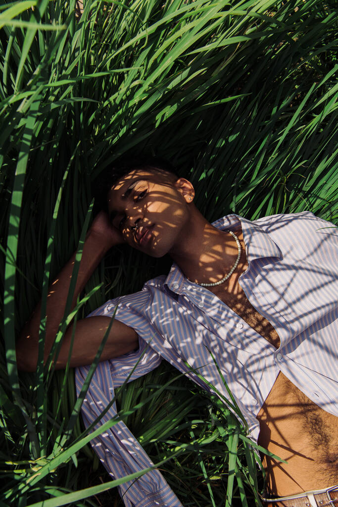 Yeşil çimenlerin üzerinde yatan mavi çizgili gömlekli Afro-Amerikan adam. - Fotoğraf, Görsel