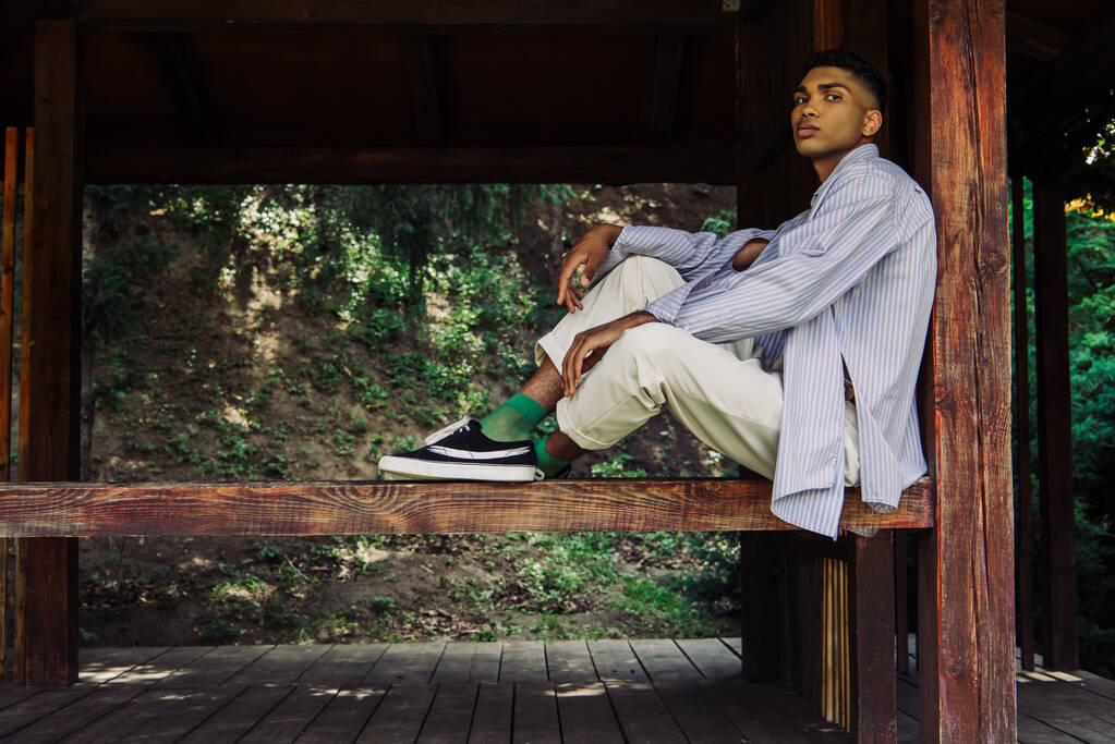 africký Američan muž v modré pruhované košili a tenisky sedí v dřevěné terase - Fotografie, Obrázek