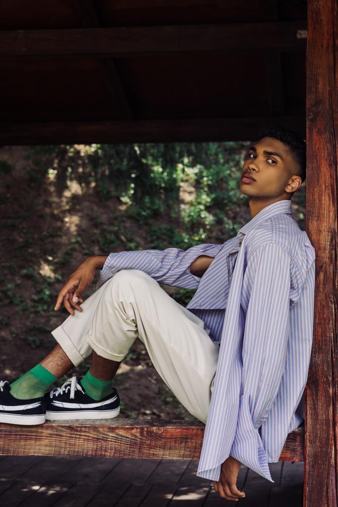 młody Afroamerykanin człowiek w niebiesko-paski koszula siedzi na drewnianym tarasie w parku - Zdjęcie, obraz