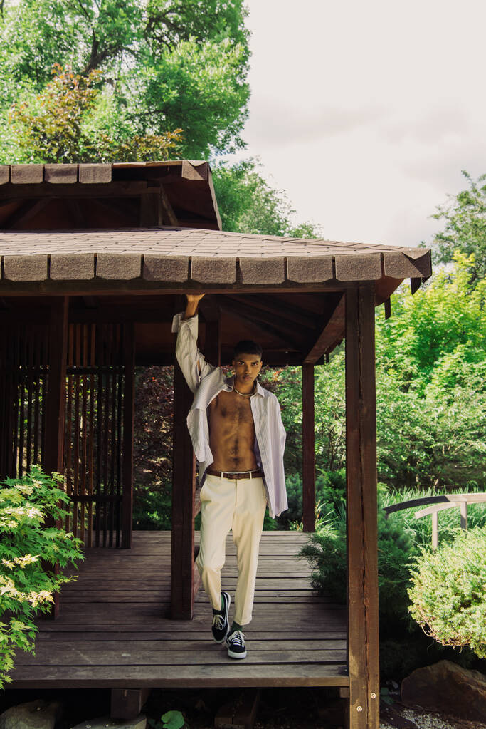 full length of african american man in trendy clothes posing in wooden patio in summer park - Fotó, kép