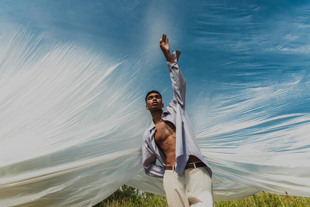 stylový africký Američan se zavřenýma očima a zvednutou rukou pod celofánovou fólií venku - Fotografie, Obrázek
