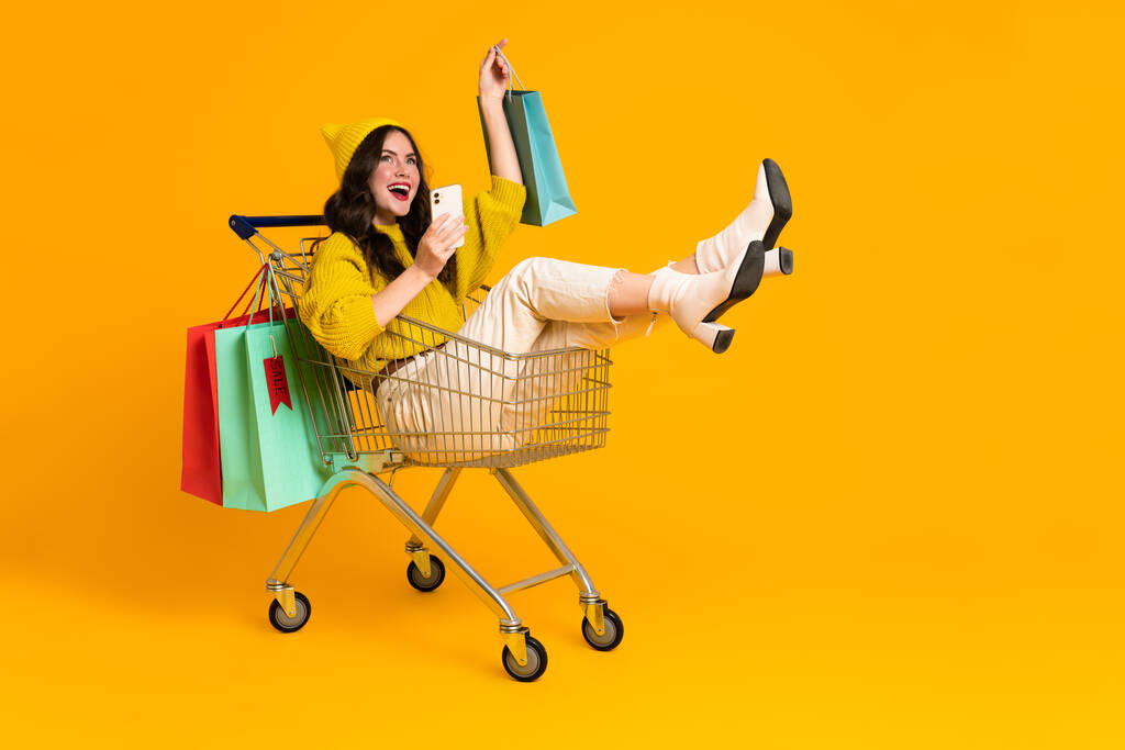 Weiße aufgeregte Frau lacht und benutzt Handy im Einkaufswagen isoliert über gelbem Hintergrund - Foto, Bild