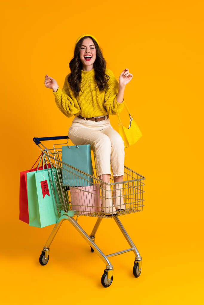 Blanke vrouw lacht terwijl ze zit in winkelwagen geïsoleerd over gele achtergrond - Foto, afbeelding